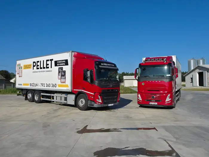 Dwie ciężarówki Volvo z zabudową PELLET na placu, w tle hala i silosy