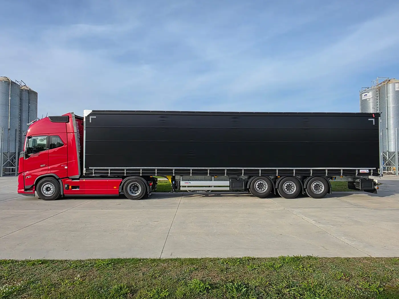 Volvo FH16 z trzyosiową naczepą kurtynową, widok z boku, w tle silosy